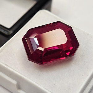 Può includere: Una gemma rettangolare rosso intenso con taglio a smeraldo, caratterizzata da molteplici sfaccettature che riflettono la luce. La pietra è posizionata su un cuscinetto espositivo bianco quadrato, con il coperchio di una scatola nera visibile.