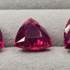 May include: Three loose, faceted gemstones in a vibrant magenta-pink color, each cut into a triangular shape. The stones feature multiple polished surfaces that reflect light, creating a brilliant appearance with sharp edges and pointed corners.