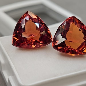Op de afbeelding: Twee driehoekige, gefacetteerde edelstenen met een warme, rood-oranje tint. De edelstenen worden gepresenteerd op een wit oppervlak, waarbij hun briljante slijpsel en interne reflecties worden benadrukt. De edelstenen zijn waarschijnlijk voor het maken van sieraden.