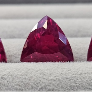 May include: Three triangular, faceted, deep red gemstones are displayed on a grey surface. The gems have a brilliant cut, reflecting light. The gems are likely rubies, showcasing a rich, saturated colour.