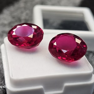 May include: Two faceted, oval-cut, deep red gemstones. The gems are displayed on a white surface, with a white box in the background. The gems are a vibrant shade of red, reflecting light.