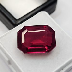 Peut inclure: Une pierre précieuse rouge foncé, taillée en rectangle, probablement un rubis, repose sur une surface blanche. La gemme est présentée sur un fond noir flou, suggérant un écrin. Les facettes de la pierre reflètent la lumière, révélant sa clarté et sa couleur.