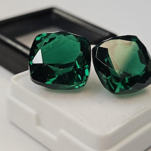 Puede incluir: Dos gemas facetadas de color verde esmeralda, exhibidas sobre una superficie blanca, con un estuche negro en el fondo. Las gemas tienen un corte cojín y reflejan la luz, mostrando su claridad y color. El entorno es sencillo, resaltando las piedras.