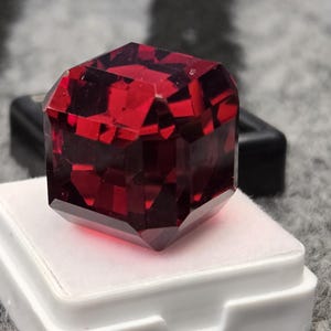 Puede incluir: Una gema facetada de color rojo intenso con forma geométrica, apoyada sobre una base cuadrada blanca. El interior de la gema refleja la luz, mostrando su estructura cristalina. El fondo es una superficie texturizada en blanco y negro.