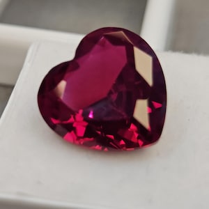 Peut inclure: Une pierre précieuse en forme de cœur, taillée à facettes, d'un magenta profond. La gemme est posée sur un fond blanc, mettant en valeur ses surfaces polies et ses reflets internes. La taille de la pierre crée de multiples facettes.