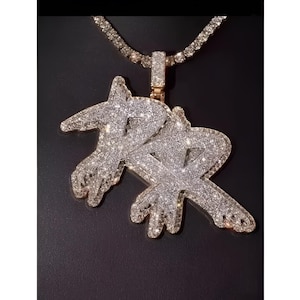 May include: A gold-toned pendant necklace with a sparkling, diamond-like encrusted design. The pendant features a stylized double-letter design, suspended from a matching chain. The necklace is displayed against a dark background.