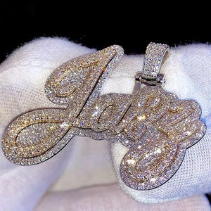 Puede incluir: Un colgante con incrustaciones de diamantes que deletrea la palabra "Love" en escritura cursiva. El colgante presenta un borde dorado y una fianza plateada. Las letras están completamente cubiertas de pequeños diamantes brillantes.
