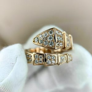 Könnte beinhalten: Ein goldfarbener Ring in Schlangenform, besetzt mit zahlreichen kleinen, klaren Edelsteinen. Der Ringkopf ist dreieckig, der Körper segmentiert. Der Ring wird vor einem weichen, neutralen Hintergrund gezeigt.