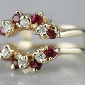 Könnte beinhalten: Ein goldfarbener Ring-Enhancer mit Doppelband-Design. Der Enhancer ist mit Clustern aus kleinen, runden, tiefroten Rubinen und funkelnden weißen Diamanten verziert. Die Edelsteine sind in einer Krappenfassung gefasst.