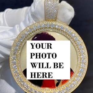 Könnte beinhalten: Ein goldfarbener Anhänger mit kreisförmigem Design, besetzt mit zahlreichen kleinen, klaren Edelsteinen. In der Mitte befindet sich ein weißes Rechteck mit dem Text "YOUR PHOTO WILL BE HERE" in fetten, schwarzen Buchstaben. Der Anhänger hat oben eine Schlaufe für eine Kette.