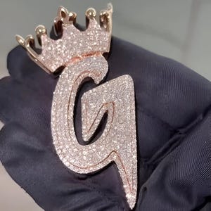 Könnte beinhalten: Ein goldfarbener Anhänger in Form des Buchstabens "G", gekrönt von einer Krone. Der Anhänger ist mit kleinen, funkelnden Steinen besetzt, die einen schillernden Effekt erzeugen. Die Krone und die Kontur des Buchstabens sind goldfarben.