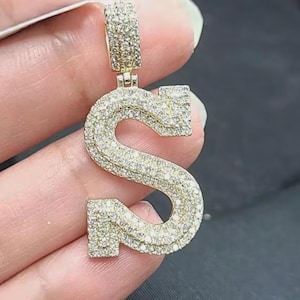 May include: A gold-colored pendant in the shape of the letter "S", encrusted with numerous small, sparkling gemstones. The pendant has a bail at the top for a chain. The gemstones are set closely together, creating a brilliant, eye-catching effect.