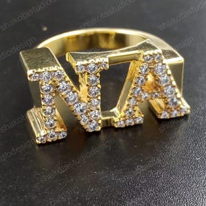 Könnte beinhalten: Ein goldfarbener Ring mit den Buchstaben "NTA", besetzt mit kleinen, klaren Edelsteinen. Der Ring hat ein dickes Band, und die Buchstaben sind erhaben und prominent, was ein auffälliges Schmuckstück ergibt. Der Ring ist wahrscheinlich aus goldfarbenem Metall.