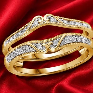 Könnte beinhalten: Ein Ring-Guard aus Gelbgold mit zwei geschwungenen Bändern, die durch dekorative Schnörkel verbunden sind. Die Bänder sind mit kleinen runden weißen Steinen im Kanal gefasst und weisen an den Kanten Milgrain-Details auf.