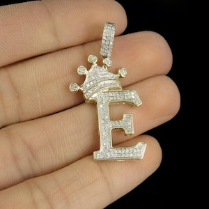 May include: A gold-toned pendant in the shape of the letter "E" topped with a crown, encrusted with small, sparkling diamonds. The pendant is held in a hand against a black background, showcasing its intricate design and craftsmanship.