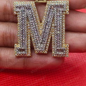 May include: A gold-toned pendant in the shape of the letter "M", encrusted with sparkling white and clear stones. The pendant has a rectangular bail and is set against a blurred background.