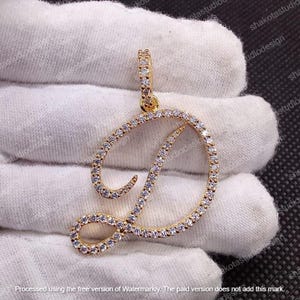 Op de afbeelding: Goudkleurige hanger in de vorm van de letter "D", bezet met kleine, heldere edelstenen. De hanger heeft een lus aan de bovenkant voor een ketting. Het ontwerp is geplaatst tegen een witte stoffen achtergrond.