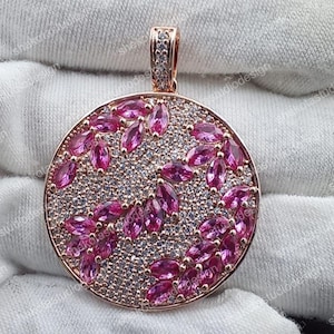 Könnte beinhalten: Roségoldfarbener Anhänger mit kreisförmigem Design. Der Anhänger ist mit kleinen weißen Steinen besetzt und weist ein Muster aus rosa, marquiseförmig geschliffenen Edelsteinen auf. Der Bügel ist ebenfalls mit kleinen weißen Steinen verziert.