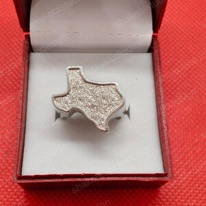 May include: A silver ring in the shape of the state of Texas, displayed in a red jewelry box. The ring has a textured surface, and the box is lined with white fabric. The ring is a unique piece of jewelry.