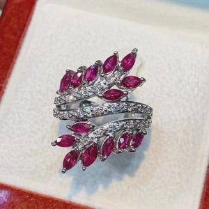 Könnte beinhalten: Ein silberfarbener Ring mit einem Bypass-Design aus zwei blattförmigen Zweigen. Jeder Zweig ist mit mehreren tiefroten Rubinen im Marquise-Schliff und kleinen, klaren Pavé-gefassten Steinen entlang der Metallbänder verziert, was ein florales Aussehen ergibt.