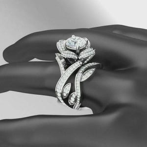 May include: A silver ring with a rose design. The ring features a large, round, clear gemstone at the center, surrounded by smaller gemstones. The band and petals are adorned with additional small gemstones, creating a sparkling effect.