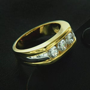 Könnte beinhalten: Ein gold-silberner Ring mit drei klaren, runden Edelsteinen in einem Kanal. Der Ring hat eine polierte Oberfläche und wird auf einer schwarzen Oberfläche präsentiert. Der Ring ist ein Schmuckstück.