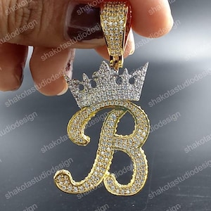 May include: A gold-colored pendant in the shape of the letter "B" with a crown on top. The pendant is encrusted with small, clear gemstones. The bail is also gold-colored and covered in gemstones. The pendant is held by a hand.