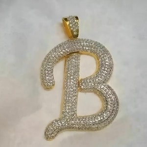 May include: A gold-toned pendant in the shape of the letter "B", encrusted with numerous small, sparkling stones. The bail is also gold and similarly adorned. The pendant is designed to be worn as jewelry, likely a necklace.