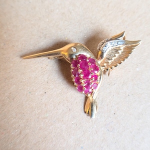 May include: A gold-toned hummingbird brooch featuring a cluster of round, vibrant pink stones on its chest, a single small clear stone set near the eye, and textured wing details with subtle silver-toned accents on the upper wing edge.