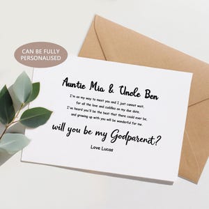 Puede incluir: Una tarjeta blanca con el texto "Auntie Mia & Uncle Ben" y "will you be my Godparent?" en escritura negra. La tarjeta va acompañada de un sobre marrón y vegetación. El texto "CAN BE FULLY PERSONALISED" está en un óvalo marrón claro.
