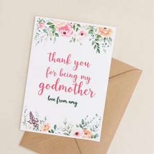Puede incluir: Una tarjeta de felicitación blanca con el texto "thank you for being my godmother" en rosa, adornada con motivos florales. Se acompaña de un sobre marrón, ideal para una ocasión especial.
