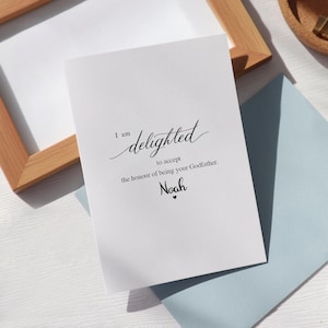 Puede incluir: Tarjeta blanca con texto negro: «Me complace aceptar el honor de ser tu padrino. Noah». La tarjeta está sobre una superficie blanca, con un sobre azul claro y un marco de madera en el fondo.