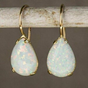 May include: A pair of gold-colored drop earrings with pear-shaped, white opal stones. The opals have a speckled appearance with hints of pink and green. The earrings are suspended from a gold-colored hook.