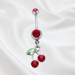 Peut inclure: Un piercing de nombril argenté avec un motif de cerise. La barre incurvée est ornée d'une gemme rouge en haut et d'une gemme rouge plus grande en bas. Un charm en forme de deux cerises rouges et d'une feuille verte est suspendu.
