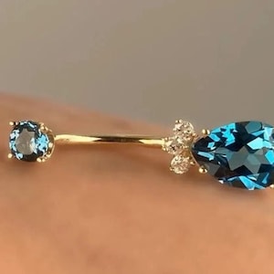 May include: A gold-colored belly button ring with two blue gemstones. One gemstone is round, and the other is a pear shape. Small clear stones are clustered near the pear-shaped gem.