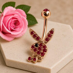 May include: A rose gold-toned navel ring featuring a rabbit head silhouette encrusted with vibrant pink crystals and a single black crystal eye. The barbell is topped with a matching rose gold-toned sphere set with a small pink crystal.