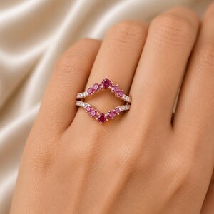 May include: A rose gold ring with a double band design. The top band features a V-shaped arrangement of pink gemstones, while the lower band is adorned with small white gemstones. The ring is set against a soft, neutral background.