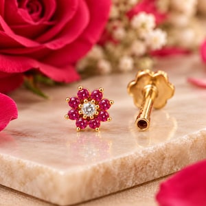 May include: A gold stud earring shaped like a flower, featuring a central clear stone surrounded by eight vibrant pink stones. The earring has a straight post and a flat, circular backing, showcasing a detailed metallic finish.