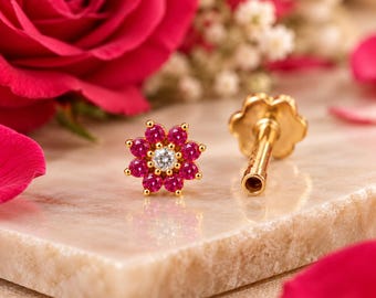May include: A gold stud earring shaped like a flower, featuring a central clear stone surrounded by eight vibrant pink stones. The earring has a straight post and a flat, circular backing, showcasing a detailed metallic finish.