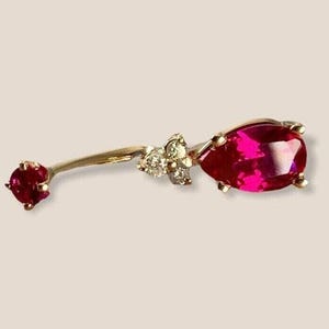 May include: A curved, gold-coloured belly button ring with a large, pear-shaped, bright pink gemstone. The ring also has a small, round, red gemstone and a cluster of small, clear gemstones.