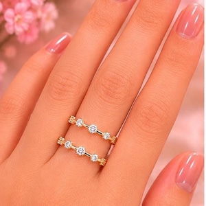 May include: Two gold-colored rings, each adorned with small, sparkling gemstones, are displayed on a person's finger. The rings have a delicate design, with the gemstones set in a row. The background is a soft pink, with hints of flowers.