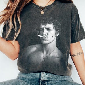 May include: Dark gray t-shirt featuring a black and white image of a person smoking a cigarette. The person has curly hair and a cross necklace. The shirt is worn with blue jeans.