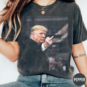 May include: Dark gray t-shirt featuring a graphic of a man in a suit giving the middle finger. The image is printed on a vintage-style tee. The shirt is worn by a person wearing blue jeans.