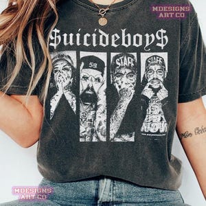 May include: Dark grey t-shirt with the band name "Suicideboys" in a gothic font. Below is a graphic of four band members in separate panels, with tattoos and hats. The shirt has a vintage, worn appearance.