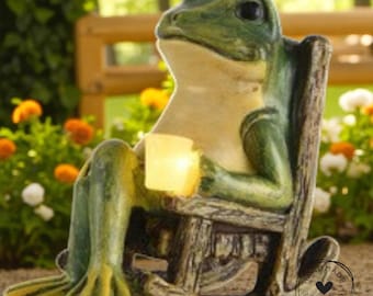 Könnte beinhalten: Eine skurrile grüne Froschstatue, die in einem hölzernen Schaukelstuhl sitzt und einen leuchtenden Würfel hält. Der Frosch hat einen hellbeige Bauch und gelb-grüne Füße. Umgeben von bunten Blumen, ideal für die Gartendekoration.