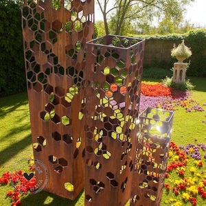 May include: Three weathered metal garden sculptures with a honeycomb pattern. The sculptures are rectangular and vary in height, with a rusty brown finish. They are set in a garden with colorful flowers and greenery.