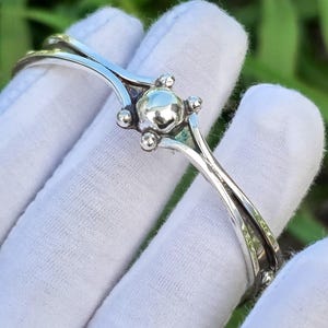 Puede incluir: Una pulsera de puño plateada con un diseño central redondeado y pequeños detalles esféricos. La pulsera presenta un diseño calado delicado con líneas curvas. La joya se muestra sobre un fondo de follaje verde.