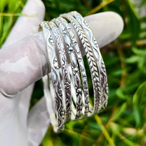 May include: A set of six silver bangles with intricate floral and leaf patterns. The bangles are displayed on a gloved finger against a backdrop of green foliage. Each bangle has a unique design, adding to the set's visual appeal.