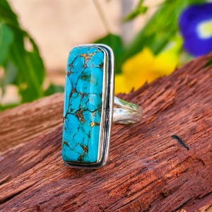 May include: A rectangular turquoise ring with gold veining set in a silver band. The turquoise stone is a vibrant blue-green color. The ring is displayed on a piece of dark brown wood.