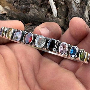 Puede incluir: Una pulsera de metal plateado que presenta una hilera de once piedras preciosas facetadas de forma ovalada y colores variados. Las piedras muestran una gama de colores que incluye rojo, azul, amarillo, morado, negro y transparente.